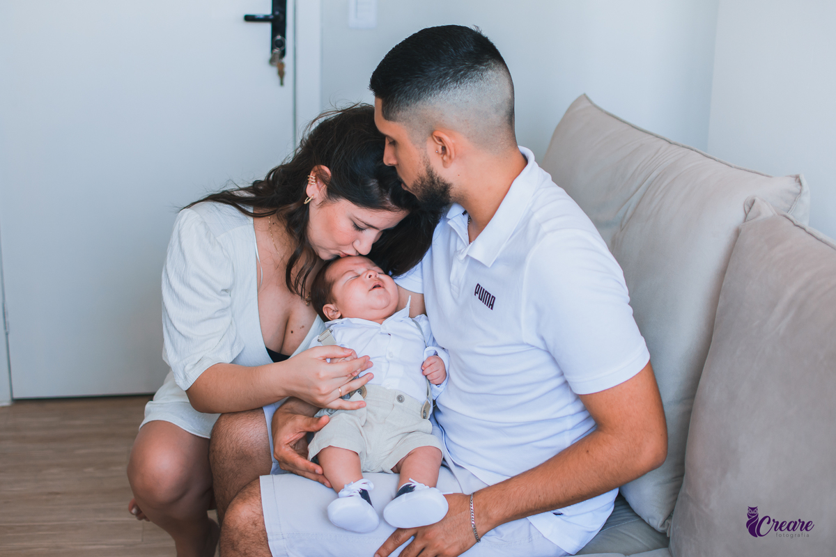 Ensaio newborn lifestyle, ensaio família. A imagem contém: um homem jovem com roupa branca, uma mulher jovem com roupa branca, um bebê com roupa branca e bermuda bege com suspensório. fotógrafo Mauá, ensaio lifestyle. 