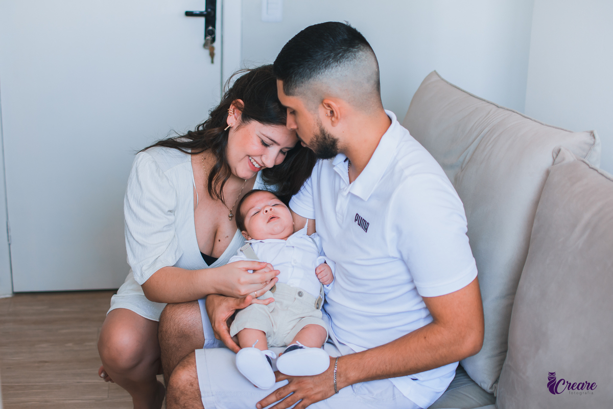 Ensaio newborn lifestyle, ensaio família. A imagem contém: um homem jovem com roupa branca, uma mulher jovem com roupa branca, um bebê com roupa branca e bermuda bege com suspensório. fotógrafo Mauá, ensaio lifestyle. 