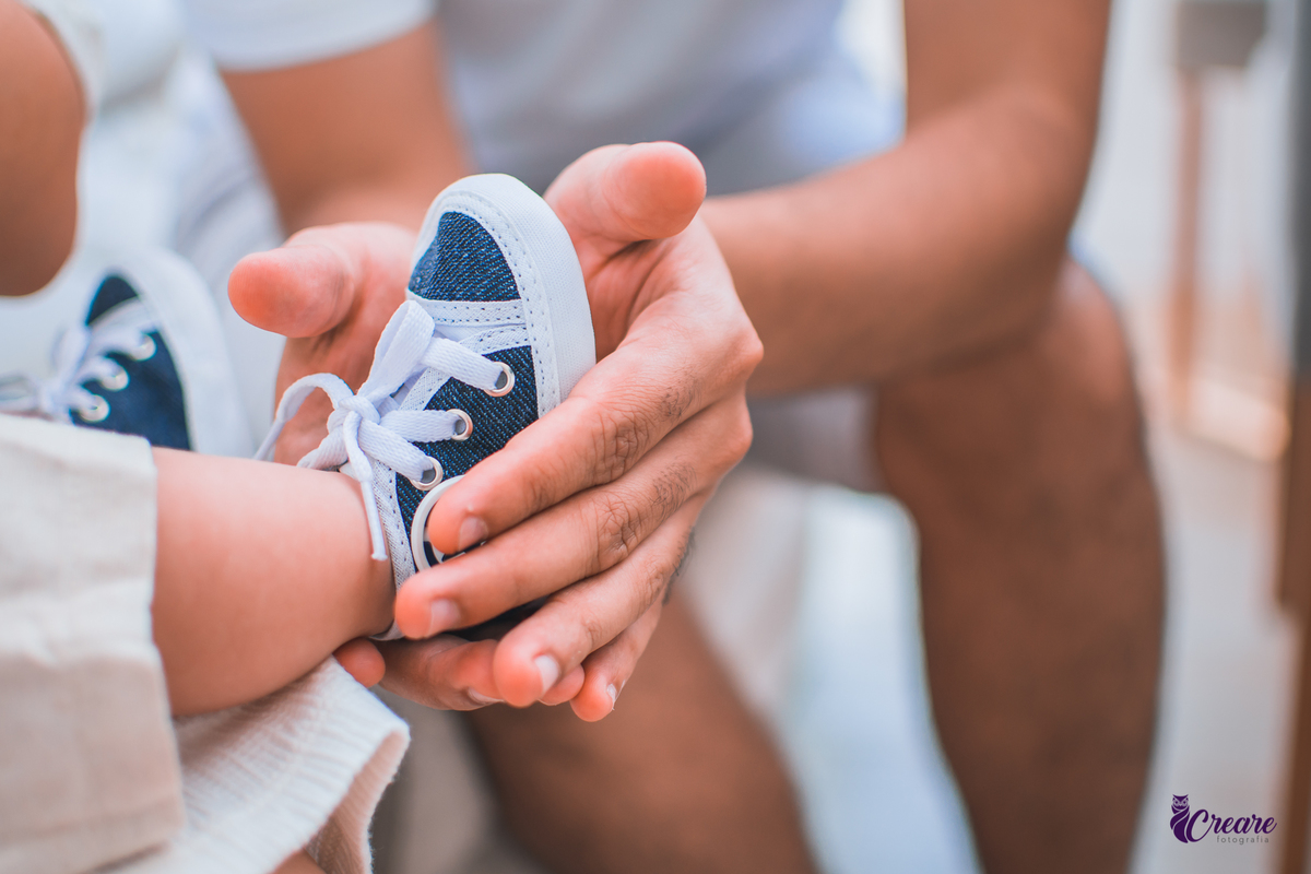Ensaio newborn lifestyle, ensaio família. A imagem contém: um homem jovem com roupa branca, uma mulher jovem com roupa branca, um bebê com roupa branca e bermuda bege com suspensório. fotógrafo Mauá, ensaio lifestyle. 