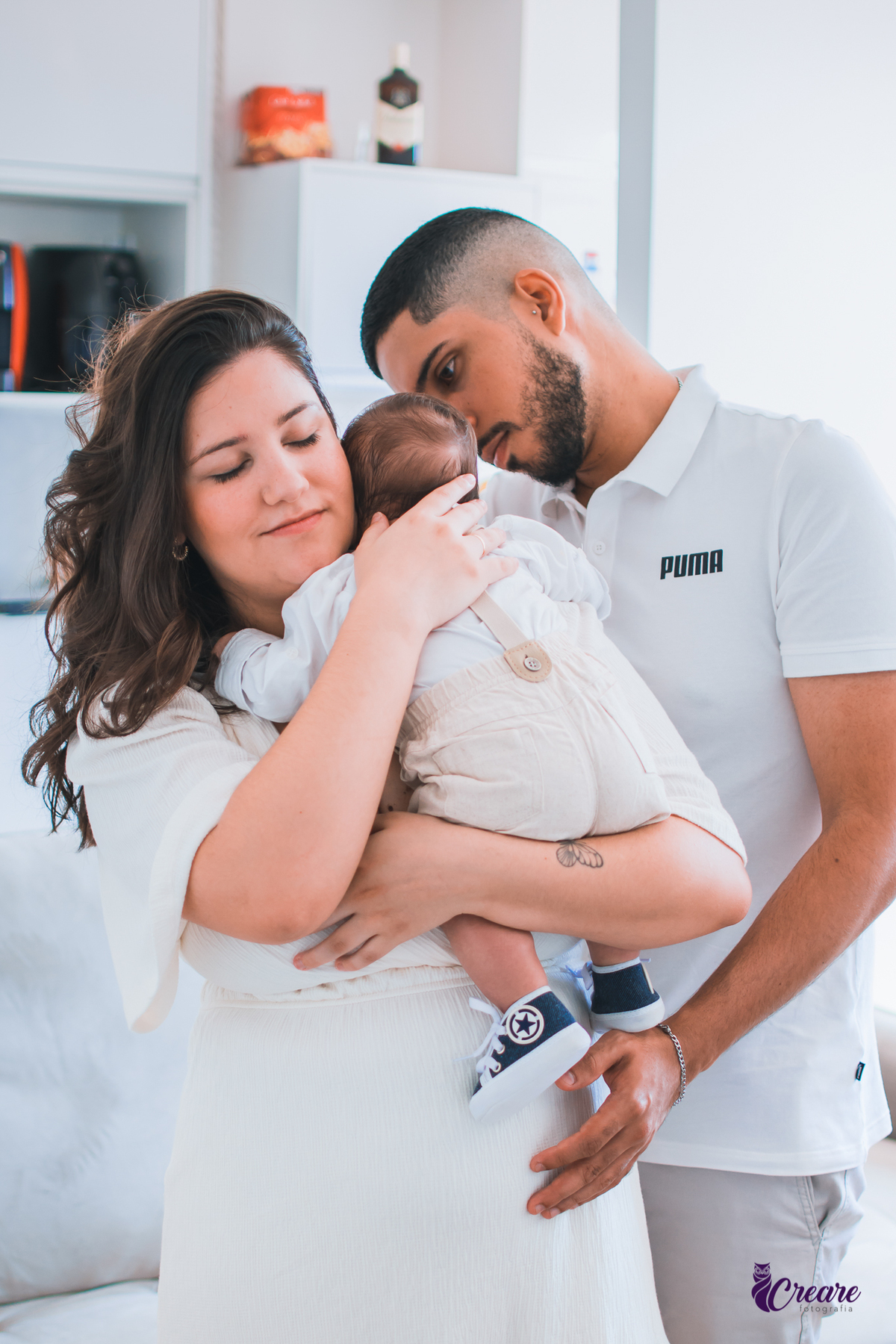 Ensaio newborn lifestyle, ensaio família. A imagem contém: um homem jovem com roupa branca, uma mulher jovem com roupa branca, um bebê com roupa branca e bermuda bege com suspensório. fotógrafo Mauá, ensaio lifestyle. 