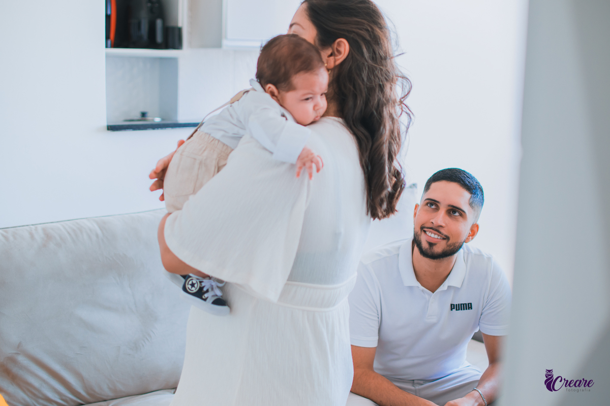 Ensaio newborn lifestyle, ensaio família. A imagem contém: um homem jovem com roupa branca, uma mulher jovem com roupa branca, um bebê com roupa branca e bermuda bege com suspensório. fotógrafo Mauá, ensaio lifestyle. 