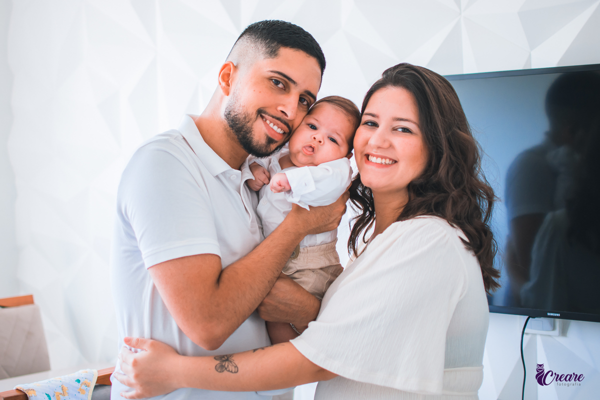 Ensaio newborn lifestyle, ensaio família. A imagem contém: um homem jovem com roupa branca, uma mulher jovem com roupa branca, um bebê com roupa branca e bermuda bege com suspensório. fotógrafo Mauá, ensaio lifestyle. 