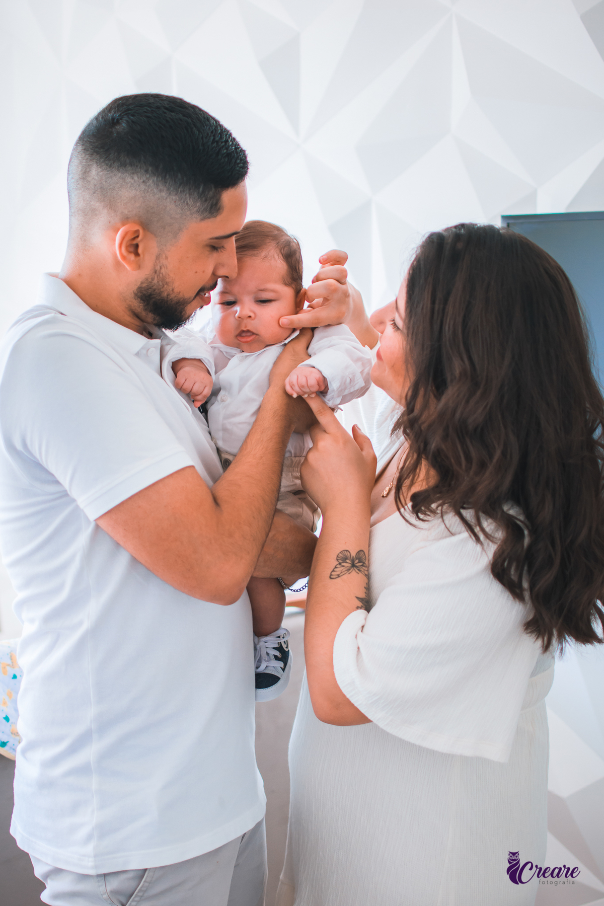 Ensaio newborn lifestyle, ensaio família. A imagem contém: um homem jovem com roupa branca, uma mulher jovem com roupa branca, um bebê com roupa branca e bermuda bege com suspensório. fotógrafo Mauá, ensaio lifestyle. 