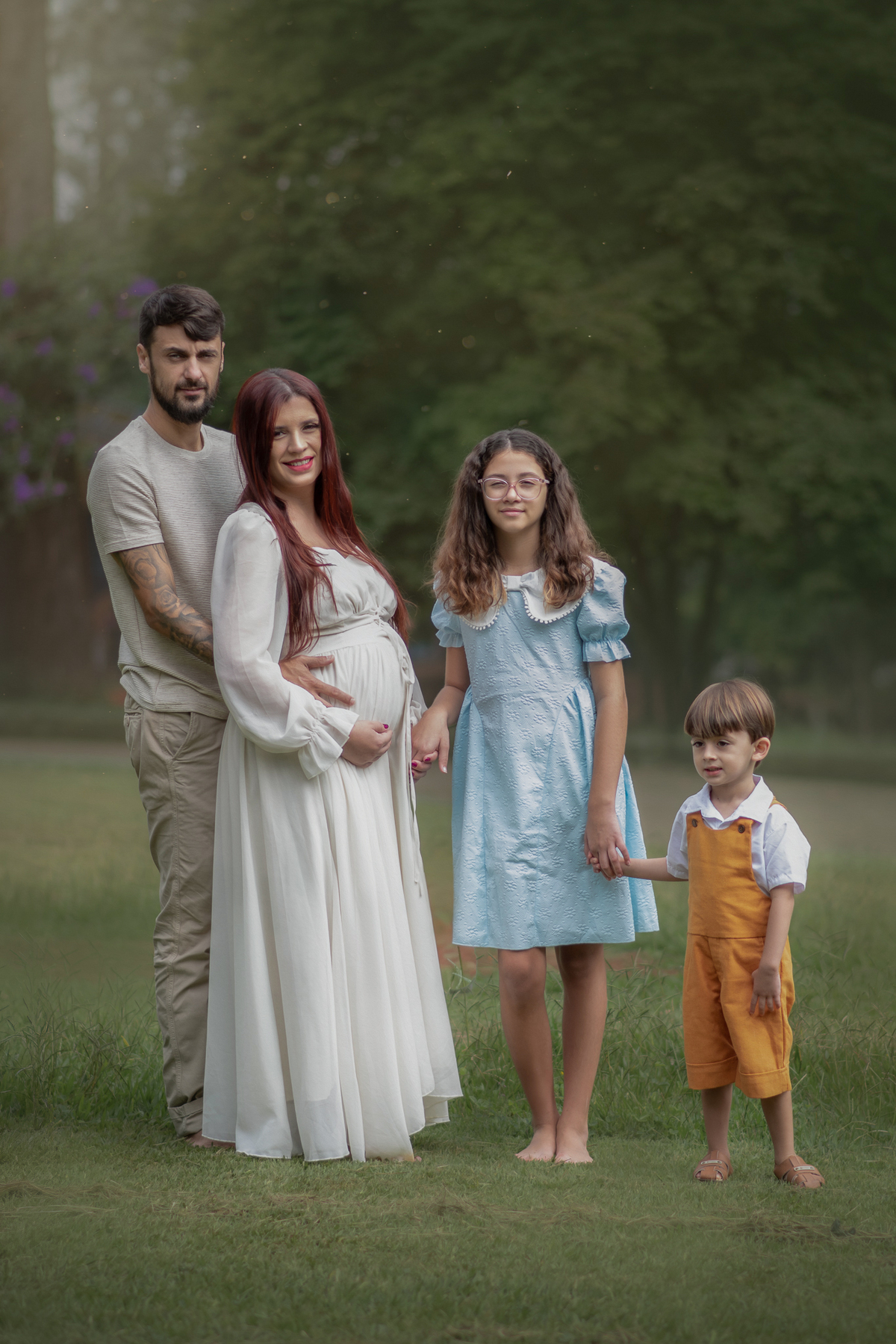 Família em ensaio ao ar livre: pai abraçando mãe grávida, adolescente de vestido azul e menino pequeno de jardineira mostarda, em pé sobre a grama com árvores ao fundo.