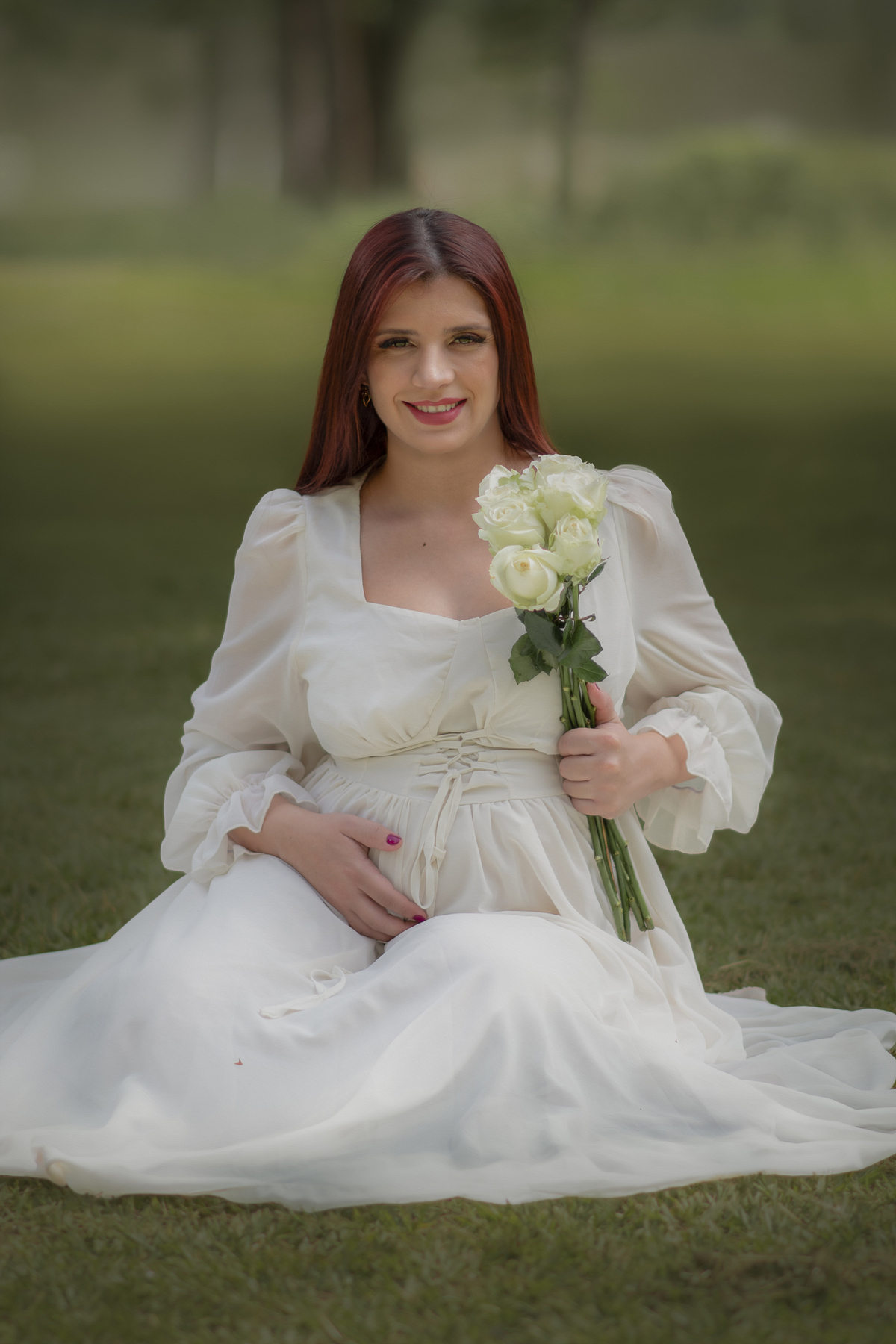 gestante de vestido branco, sentanda segurando flores brancas em um parque durante o ensaio fotográfico.