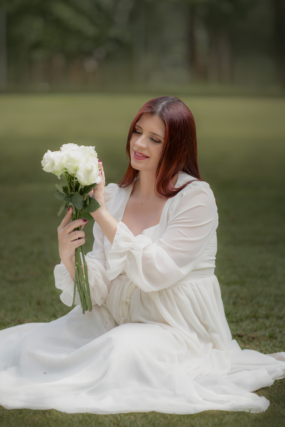 gestante de vestido branco, sentanda na grama observando flores brancas no Parque Chácara Silvestre durante o ensaio fotográfico.