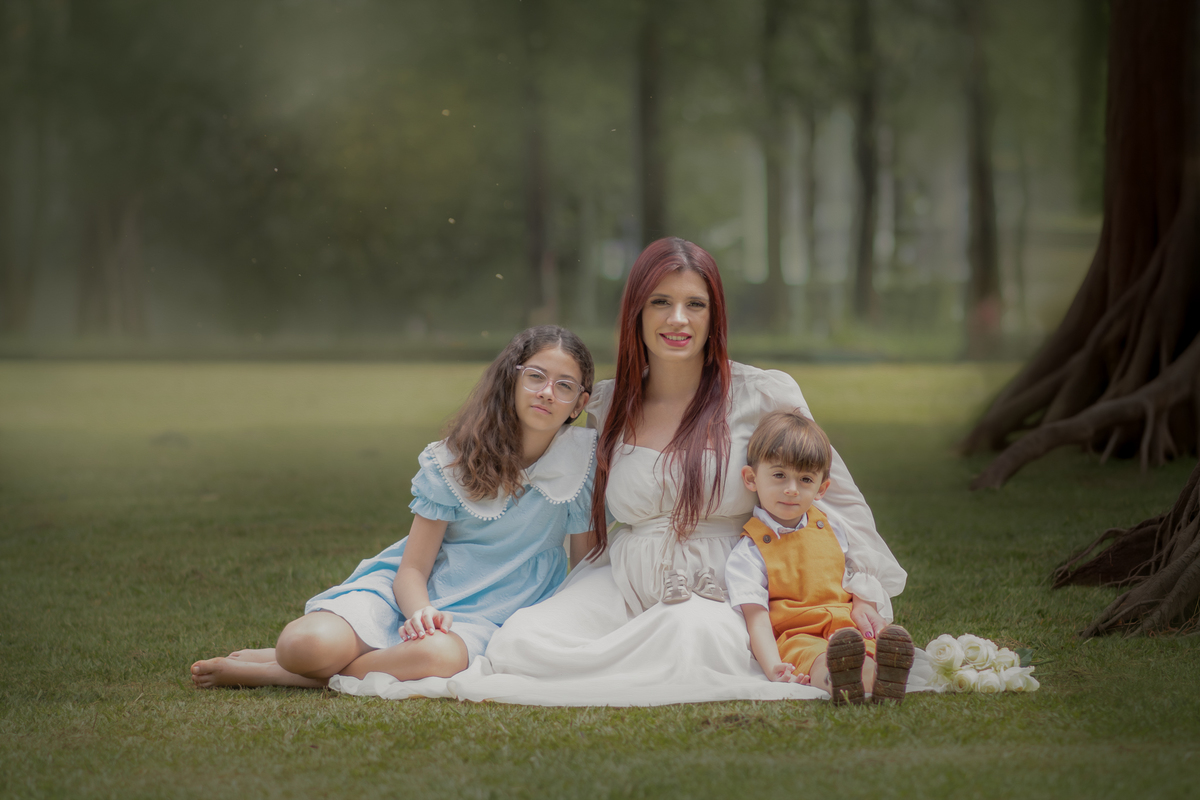 uma mãe gestante com uma adolesccente de vestido azul e um menino de jardineira mostarda, sentados na grama do Parque Chácara Silvestre durante o ensaio fotográfico de gestante.