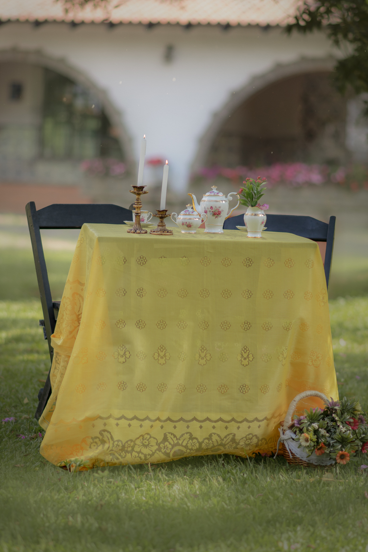 Cenário de ensaio fotográfico de dia das mães ao ar livre. Uma mesa com toalha amarela e aparelhos de chá com dois castiçais com velas.