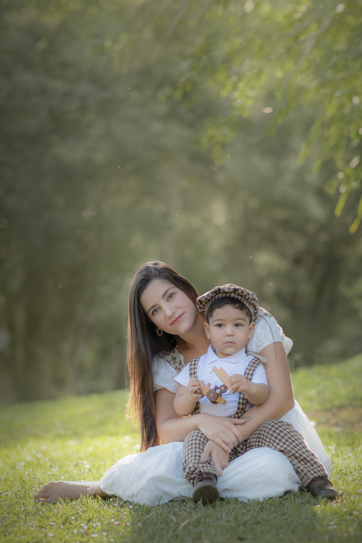 Uma mulher sentada na grama com seu filho no colo, com uma luz de fim de tarde envolvendo os dois, durante o ensaio de dia das mães ao ar livre.