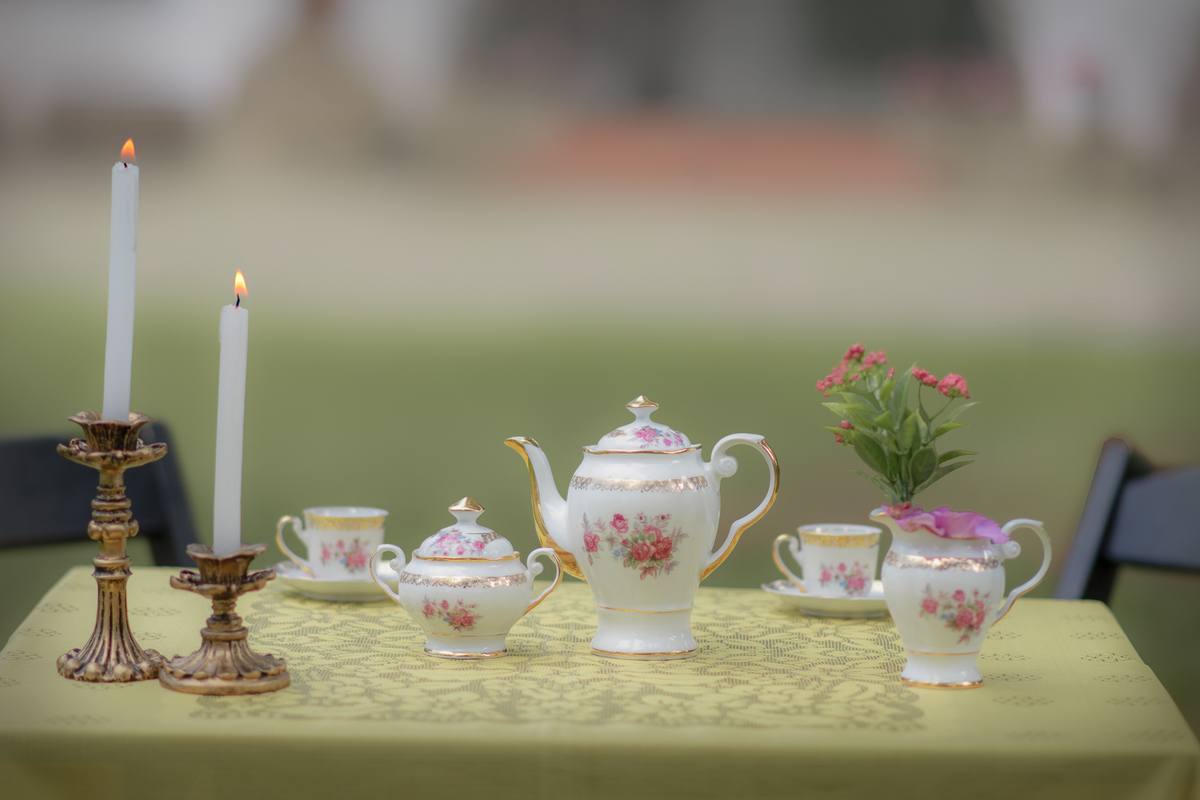 Cenário de ensaio fotográfico de dia das mães ao ar livre. Uma mesa com toalha amarela e aparelhos de chá com dois castiçais com velas.