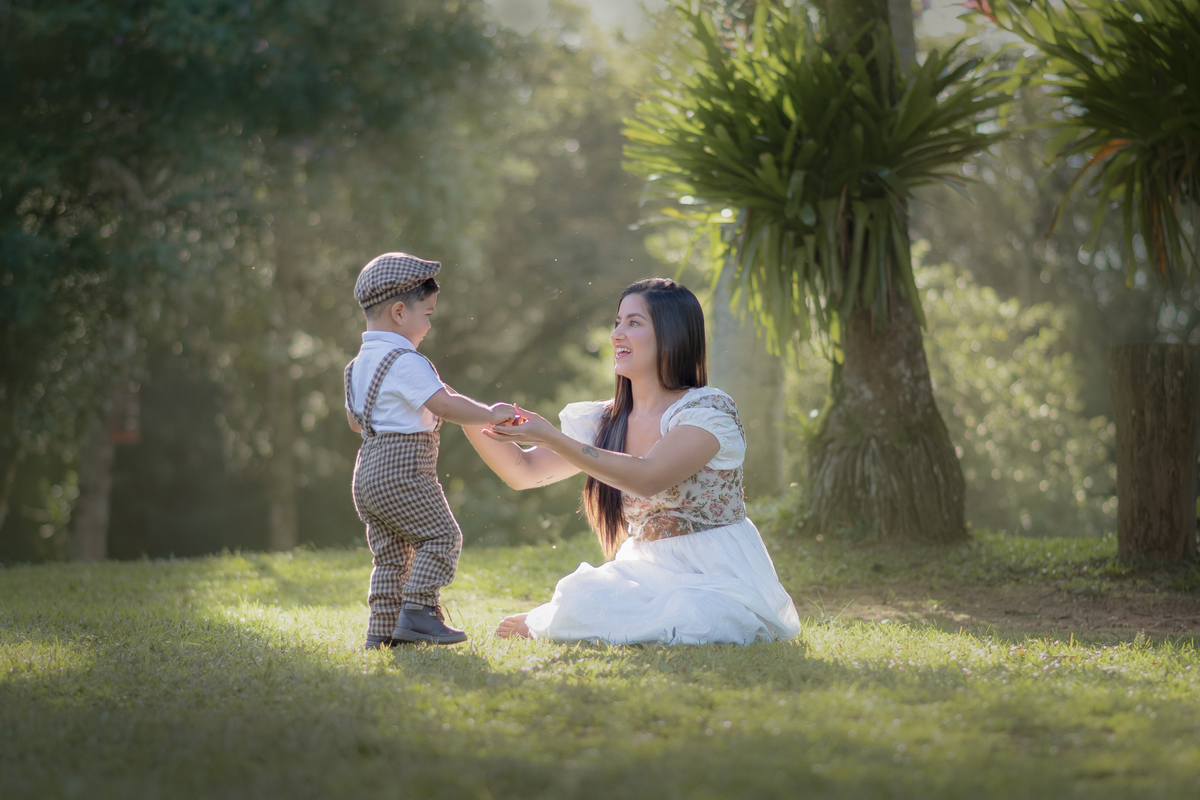 Uma mulher sentada na grama com seu filho a frente, os dois vestidos com roupas camponesas, durante o ensaio fotográfico de dia das mães.