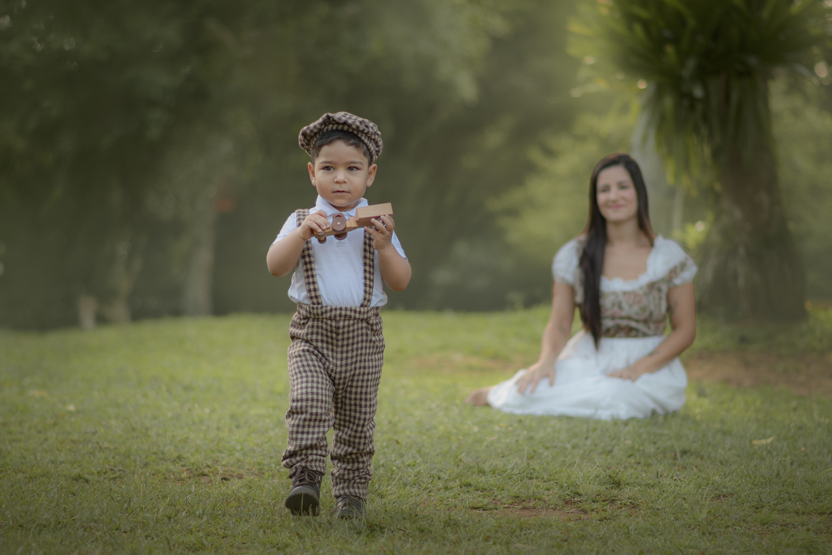 Um menino vestido com roupas campenesas, caminhando com um carrinho de madeira na mão, enquanto sua mãe o observa, durante o ensaio fotográfico de dia das mães no parque.