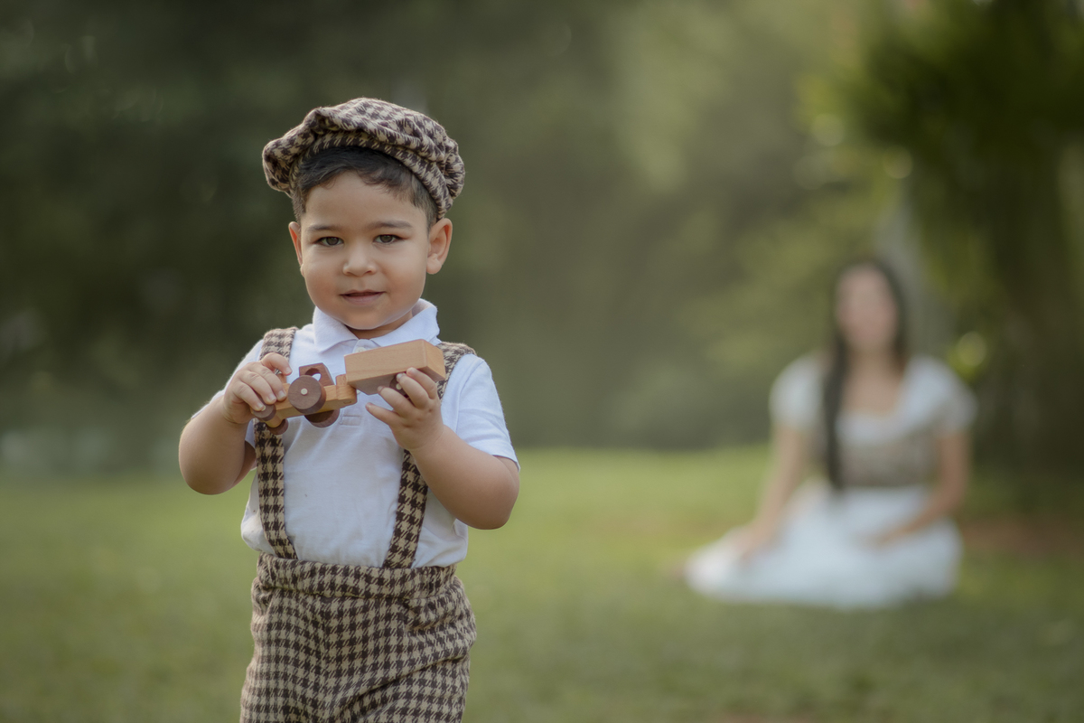 Um menino vestido com roupas campenesas, caminhando com um carrinho de madeira na mão, enquanto sua mãe o observa, durante o ensaio fotográfico de dia das mães no parque.