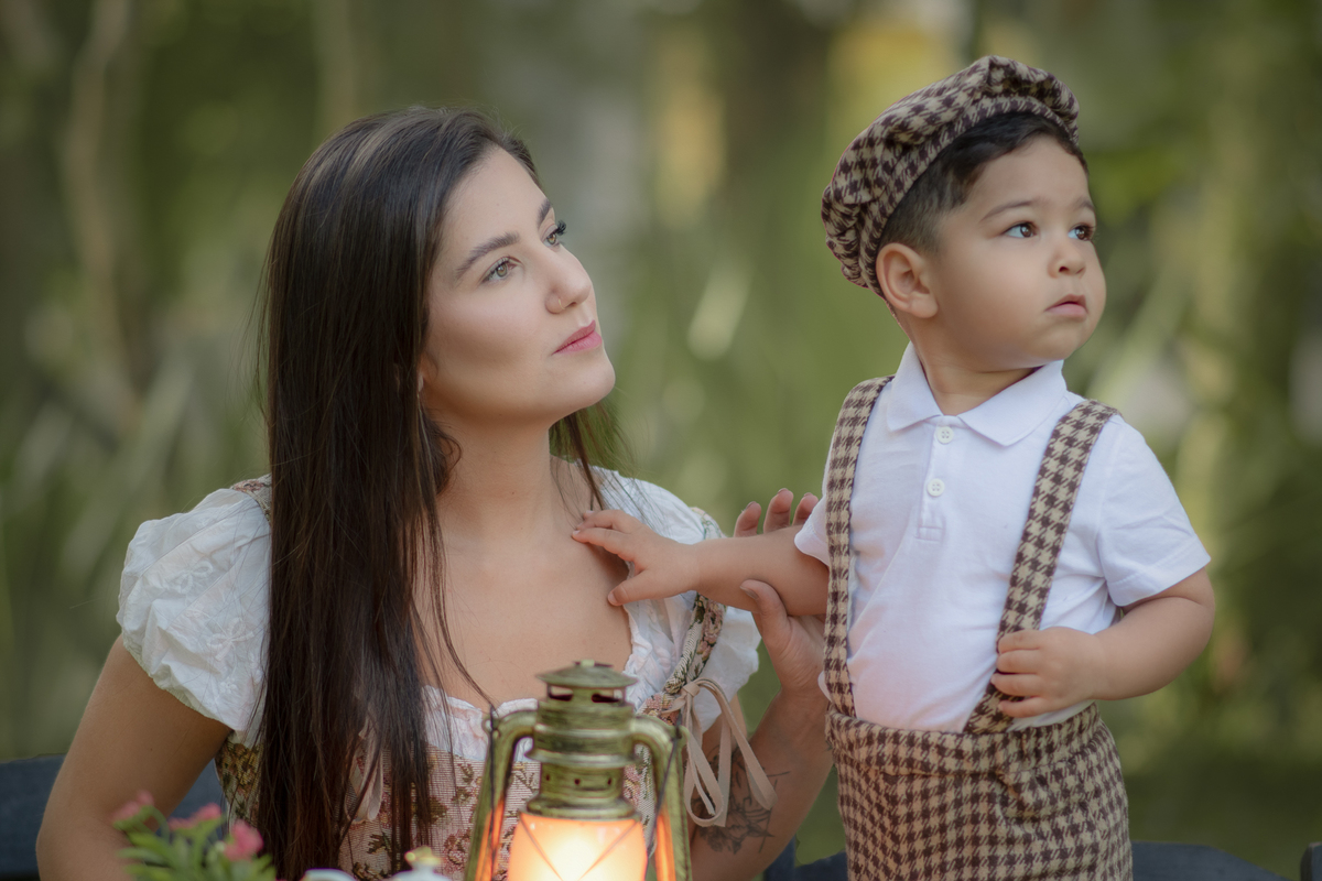 Mãe e filho sentados em uma messa em um parque, durante um ensaio fotográfico ao ar livre, os dois olhandopara o lado.
