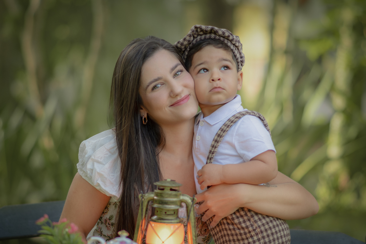 Mãe e filho sentados em uma messa em um parque, durante um ensaio fotográfico ao ar livre, o filho com o rosto encostado na mãe.