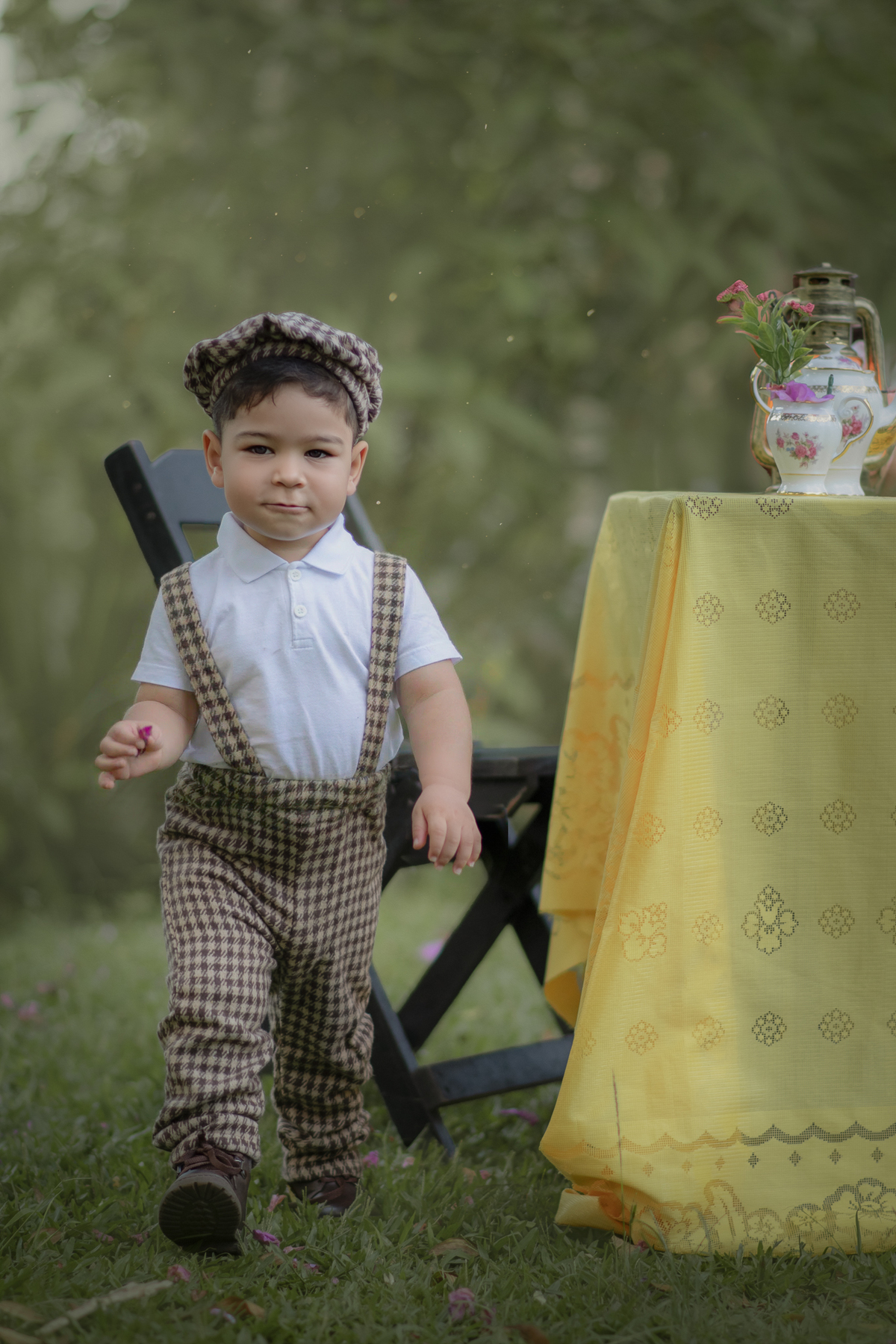 Um menino, ao lado de uma mesa com toalha amarela, vestido com roupa camponês brincando em um parque,  durante o ensaio fotográfico de dia das mães.