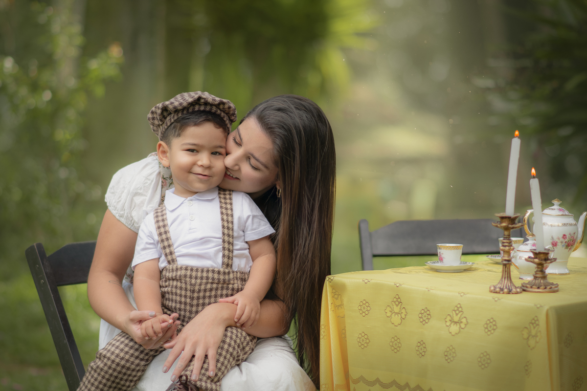 Uma mãe e seu filho no colo em Uma mesa com toalha amarela com aparelhos de chá com dois castiçais com velas. O cenário é parte de um ensaio fotográfico ao ar livre.