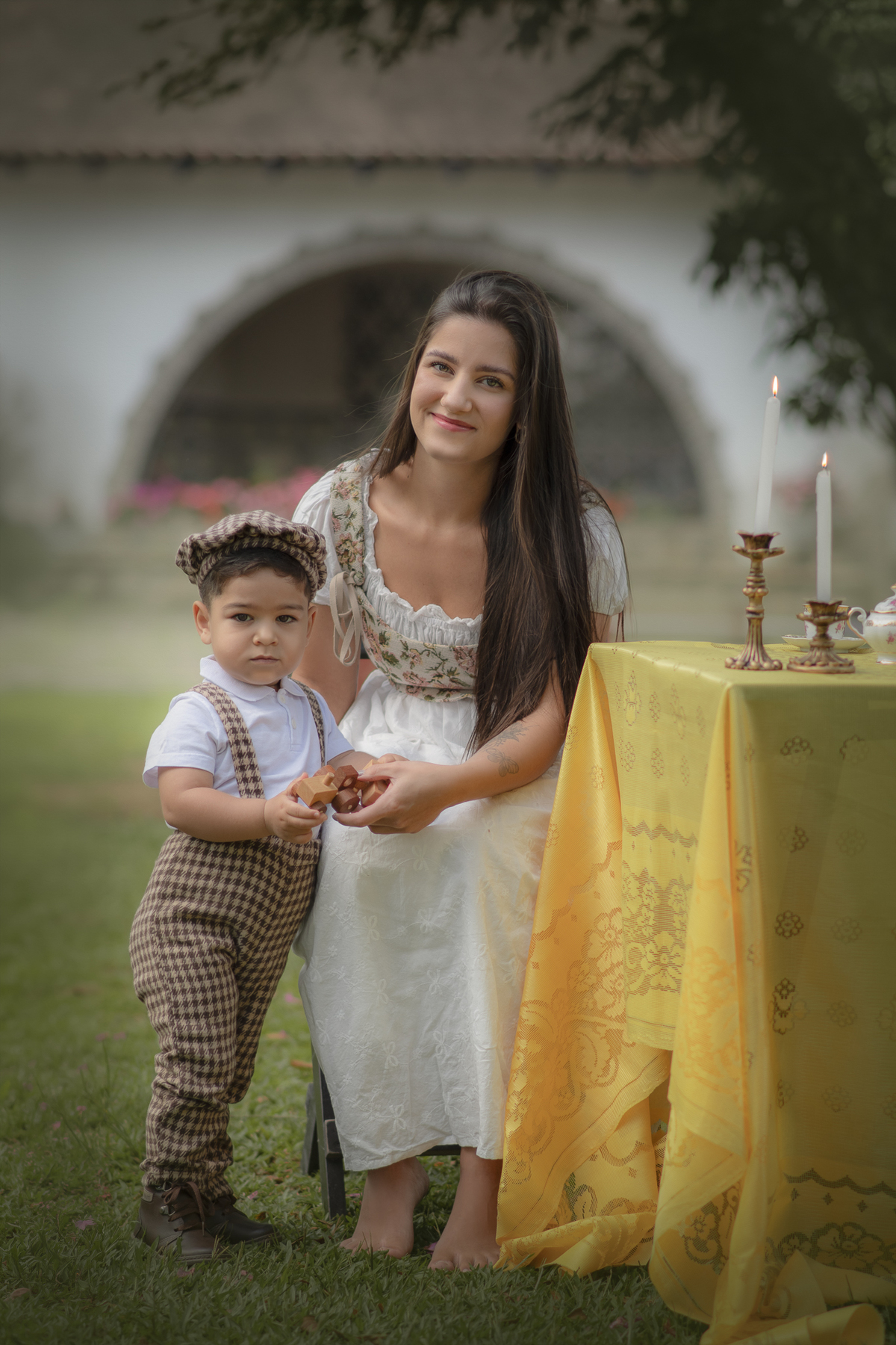 Uma mãe e seu filho em Uma mesa com toalha amarela com aparelhos de chá com dois castiçais com velas.