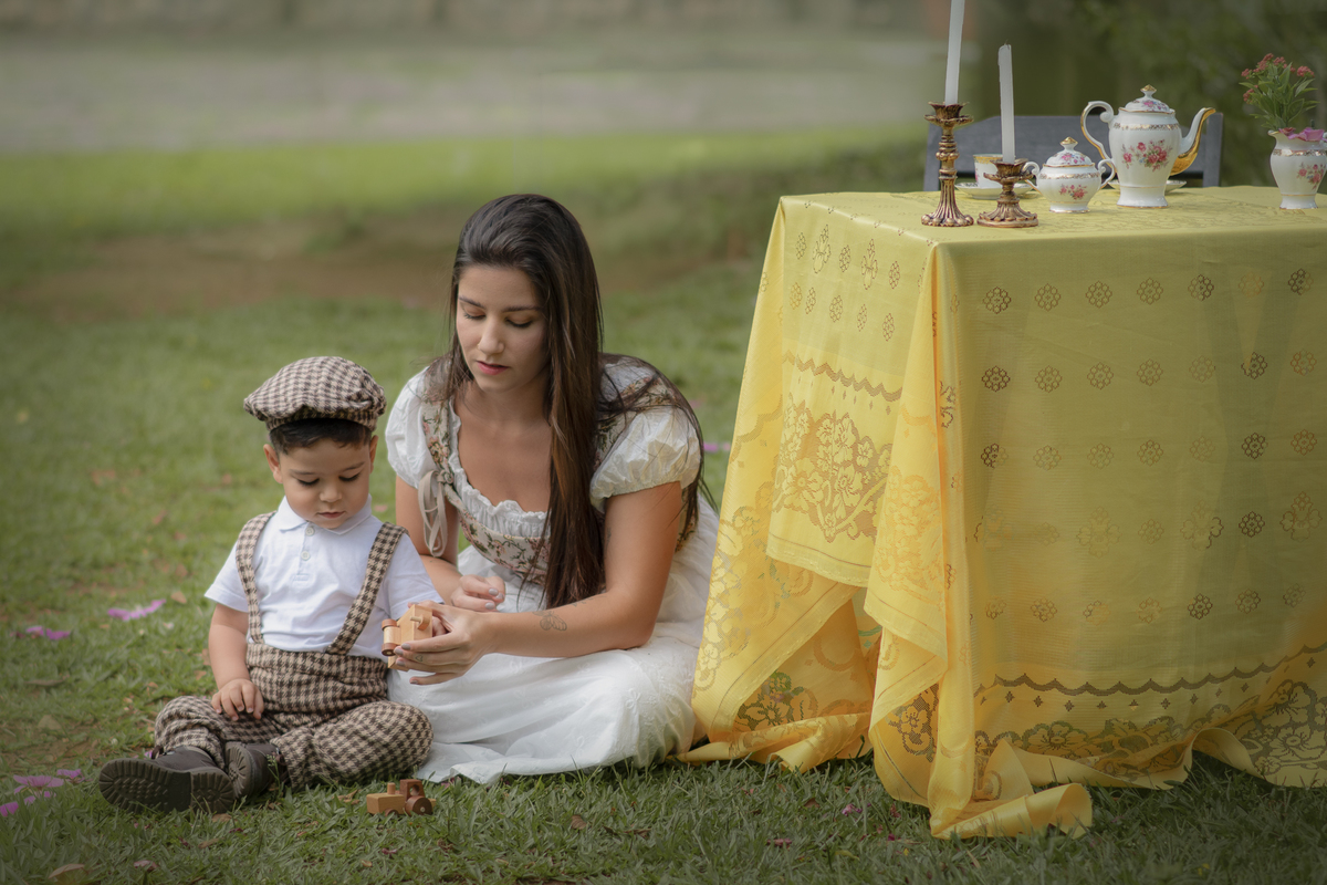 Mulher sentada na grama brincando com o filho com um carrinho de madeira, ao seu lado, uma mesa com toalha amarela preparada para um ensaio de dia das mães.