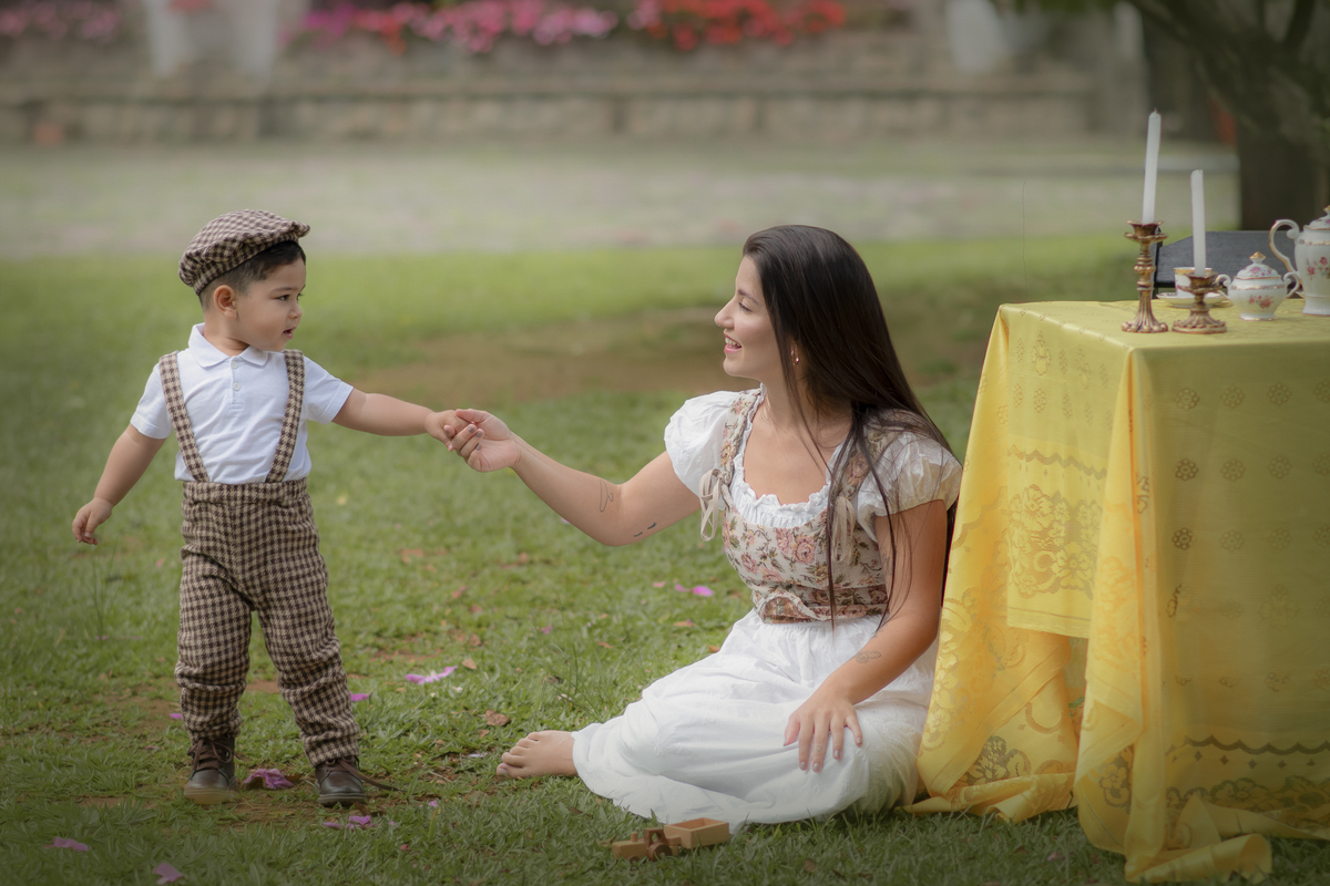lher sentada na grama segurando a mão do filho que está em pé ao seu lado, próximos a uma mesa com toalha amarela preparada para um ensaio de dia das mães.