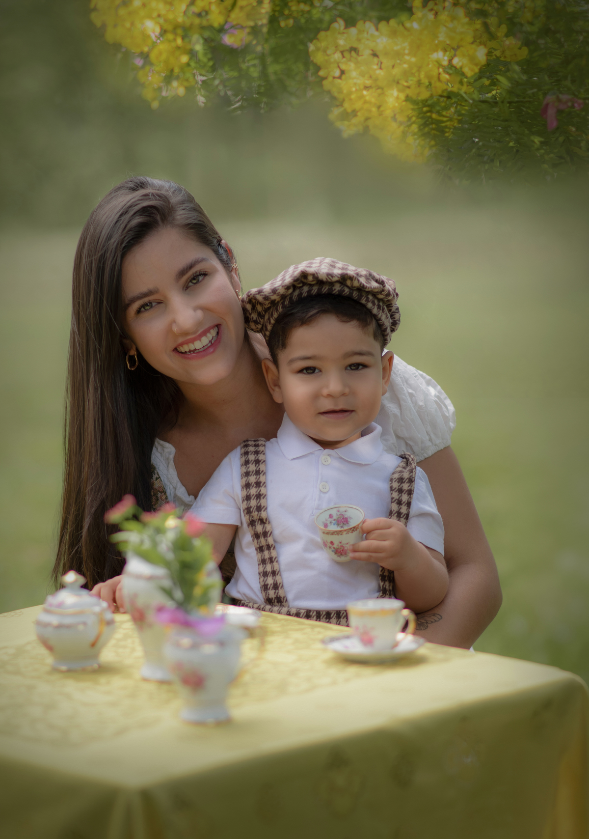 Uma mãe e seu filho no colo em Uma mesa com toalha amarela com aparelhos de chá com dois castiçais com velas. O cenário é parte de um ensaio fotográfico ao ar livre.
