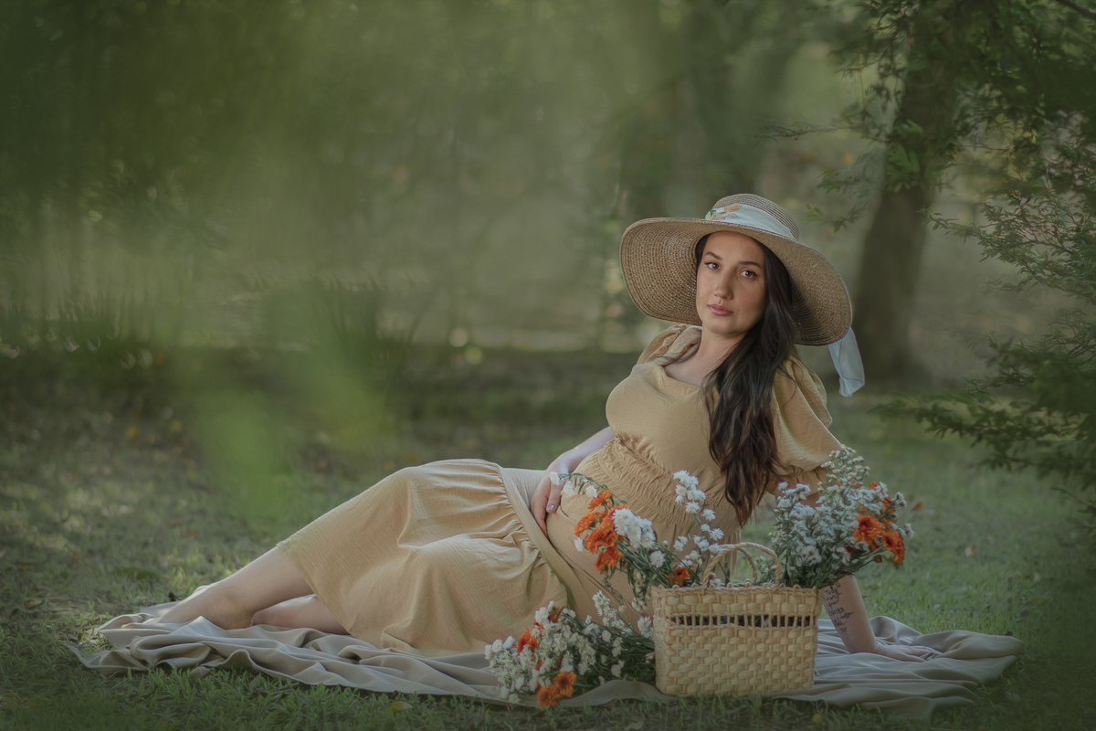 uma mulher sentada ao chão de um parque com muito verde em volta, durante um ensaio fotográfico de gestante. Ela está usando um chapéu e a sua frente tem uma cesta com flores.