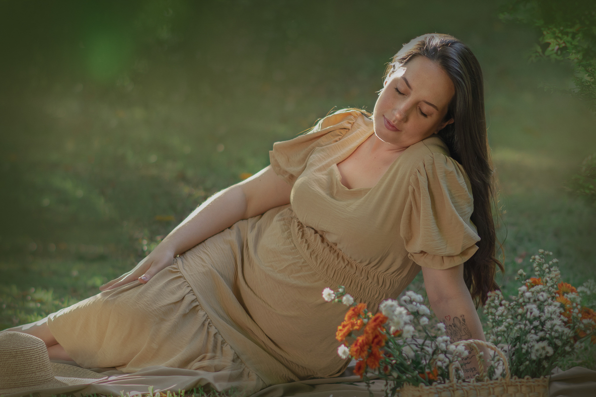 uma mulher sentada ao chão de um parque com muito verde em volta, durante um ensaio fotográfico de gestante e a sua frente tem uma cesta com flores.