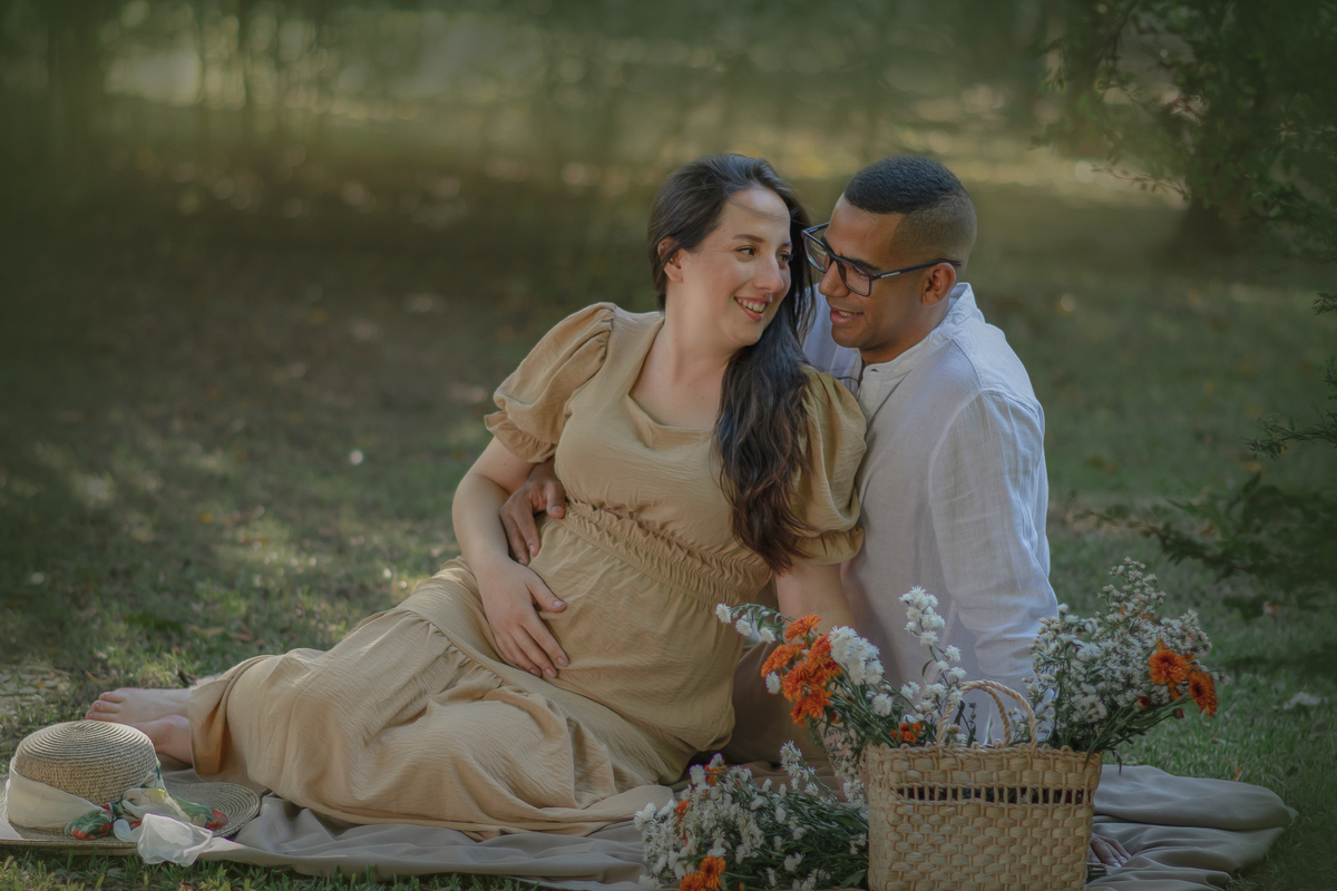 um casal sentados ao chão de um parque com muito verde em volta, durante um ensaio fotográfico de gestante e a sua frente tem uma cesta com flores.