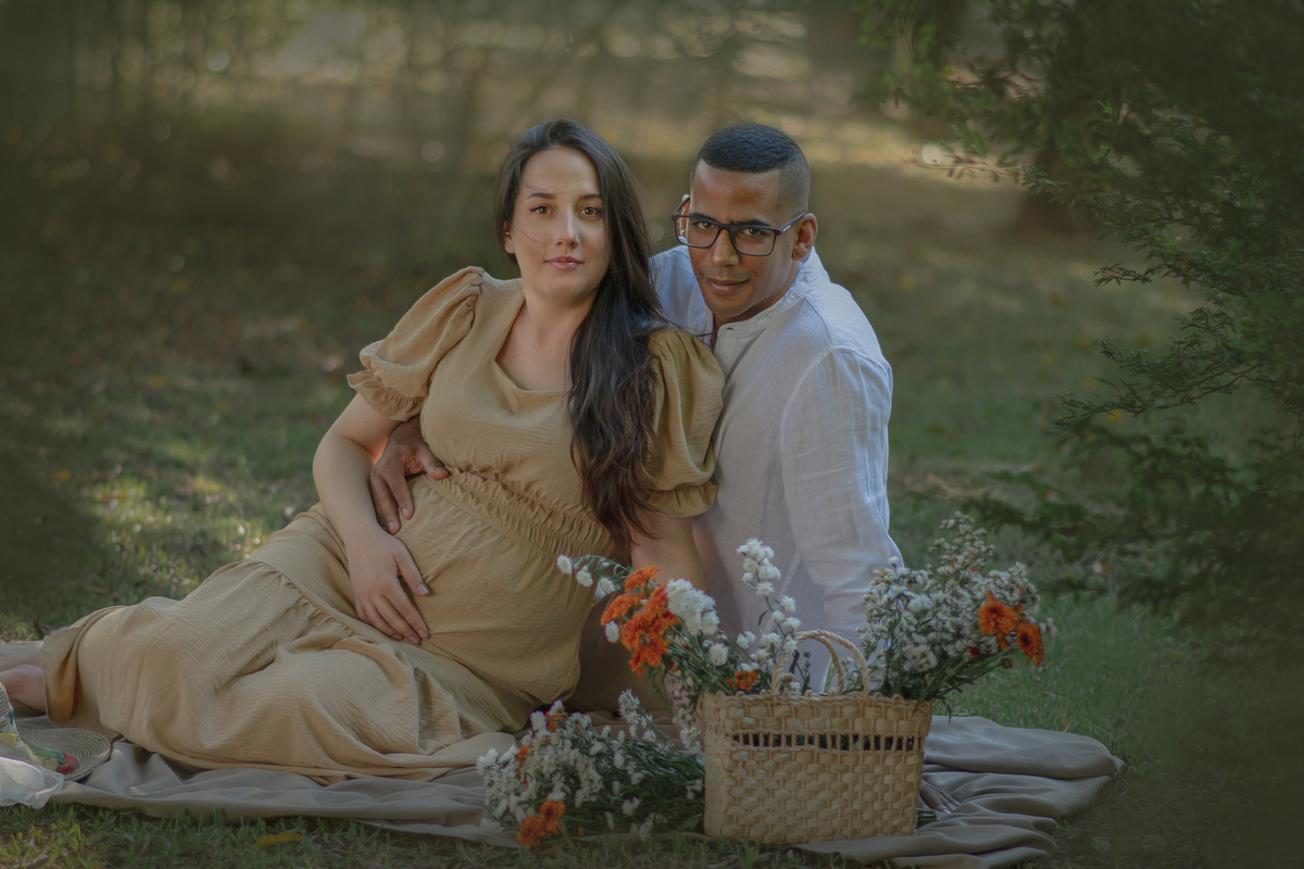 um casal sentados ao chão de um parque com muito verde em volta, durante um ensaio fotográfico de gestante e a sua frente tem uma cesta com flores.