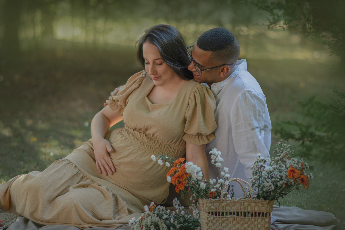 um casal sentados ao chão de um parque com muito verde em volta, durante um ensaio fotográfico de gestante e a sua frente tem uma cesta com flores.