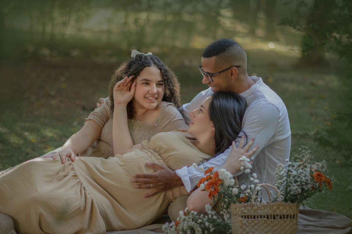 um casal sentados ao chão de um parque com muito verde em volta, durante um ensaio fotográfico de gestante e a sua frente tem uma cesta com flores.
