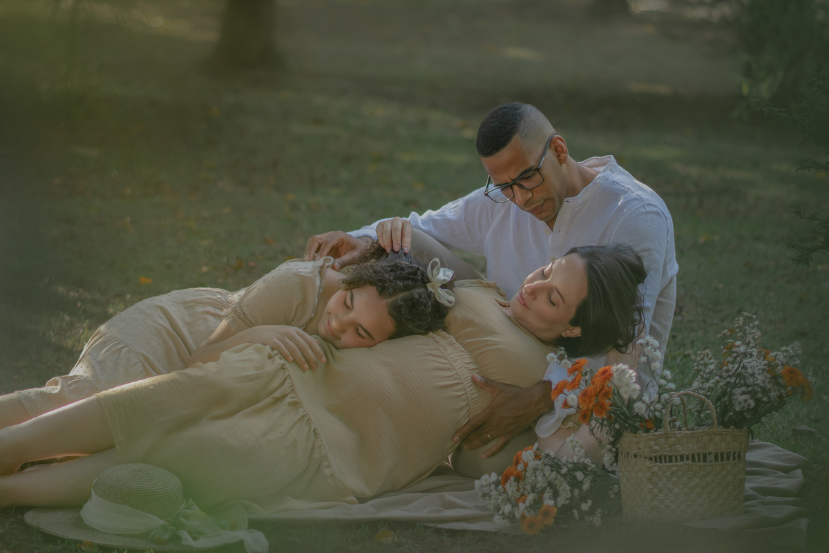 um casal e sua filha sentados ao chão de um parque com muito verde em volta, durante um ensaio fotográfico de gestante e a sua frente tem uma cesta com flores.