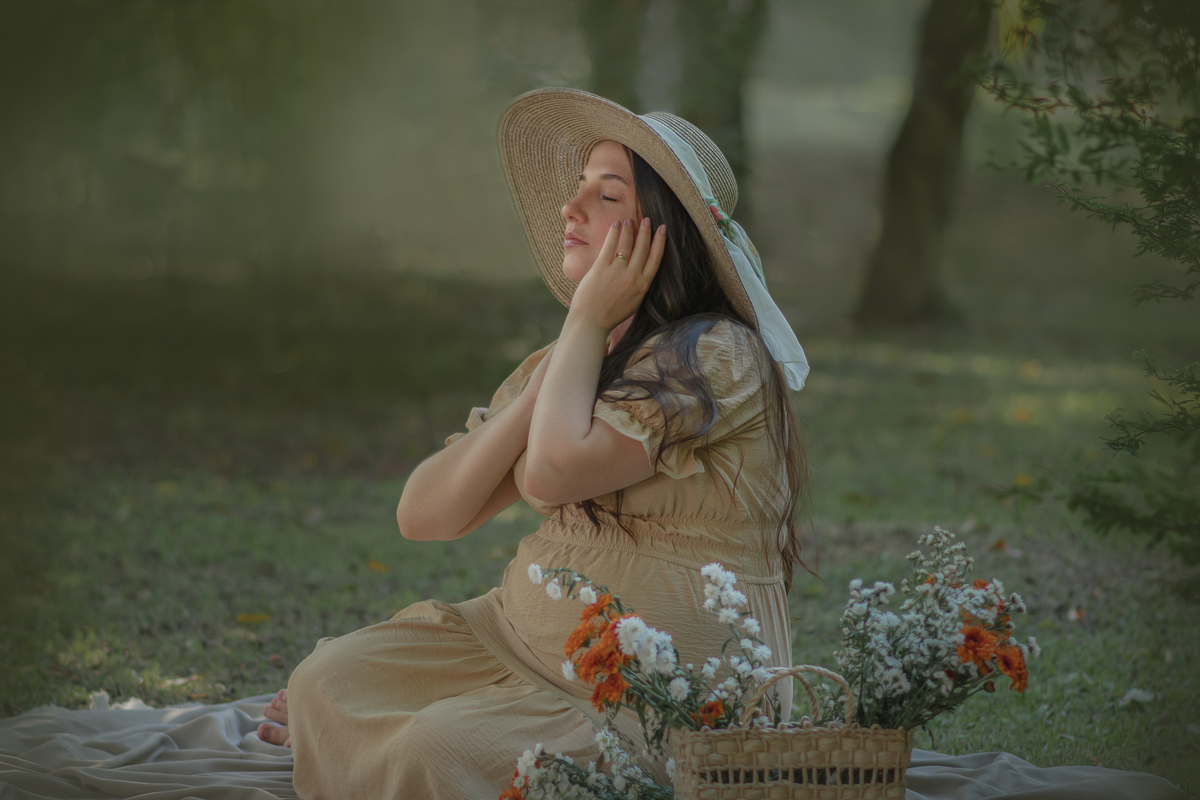 uma mulher sentada ao chão de um parque com muito verde em volta, durante um ensaio fotográfico de gestante. Ela está usando um chapéu e a sua frente tem uma cesta com flores.