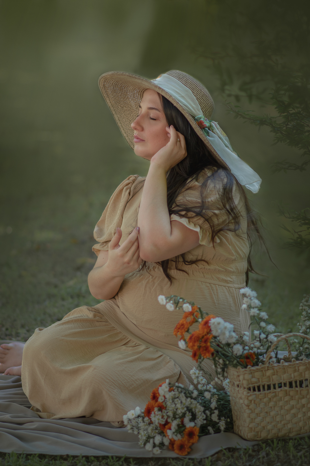 uma mulher sentada ao chão de um parque com muito verde em volta, durante um ensaio fotográfico de gestante. Ela está usando um chapéu e a sua frente tem uma cesta com flores.