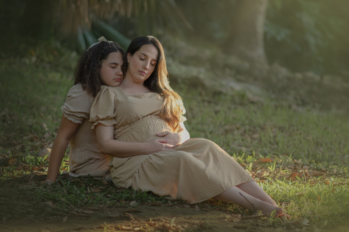 Uma mulher gestante abraçada com sua filha adolescente, com  o sol batendo em seus cabelos, durante o ensaio fotográfico no Parque Chico mendes.