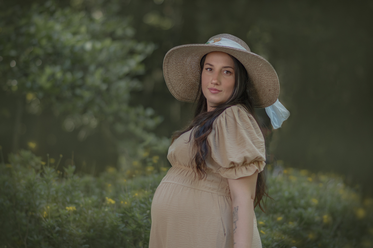 uma mulher em um parque com muito verde em volta, durante um ensaio fotográfico de gestante. Ela está usando um chapéu e carregado uma cesta de flores. 