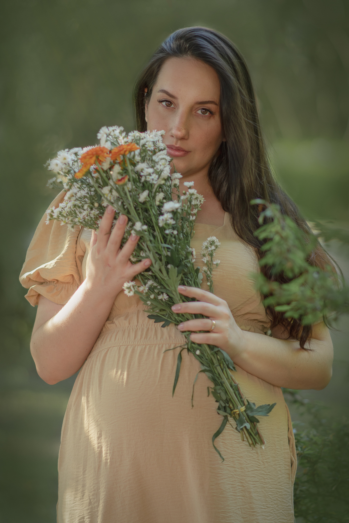 uma mulher sentada ao chão de uma parque com muito verde em volta, durante um ensaifo fotográfico de gestante. Ela está usando um chapéu e a sua frente tem uma cesta com flores.