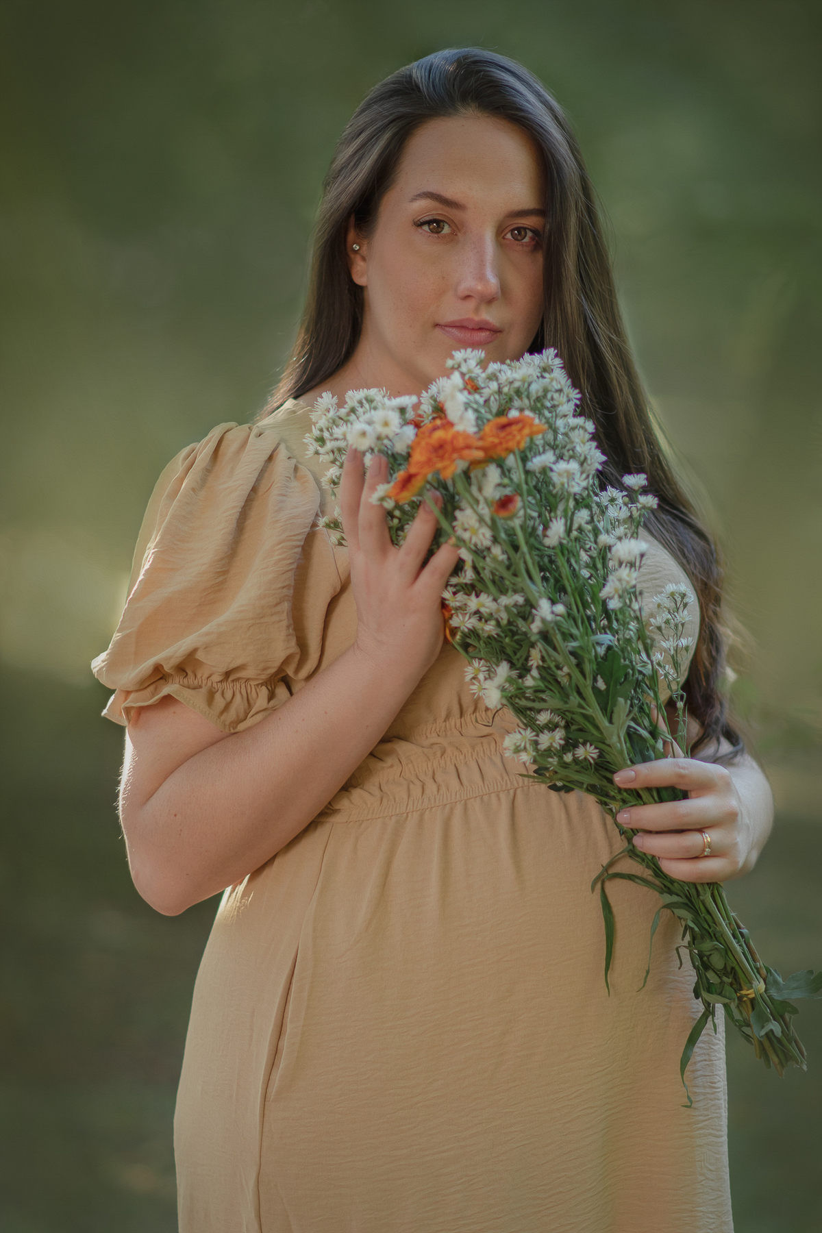 uma mulher sentada ao chão de um parque com muito verde em volta, durante um ensaio fotográfico de gestante e a sua frente tem uma cesta com flores.
