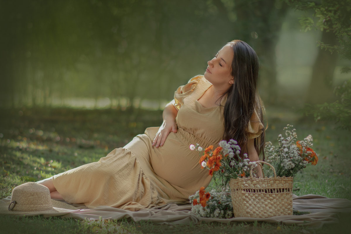uma mulher sentada ao chão de um parque com muito verde em volta, durante um ensaio fotográfico de gestante e a sua frente tem uma cesta com flores.