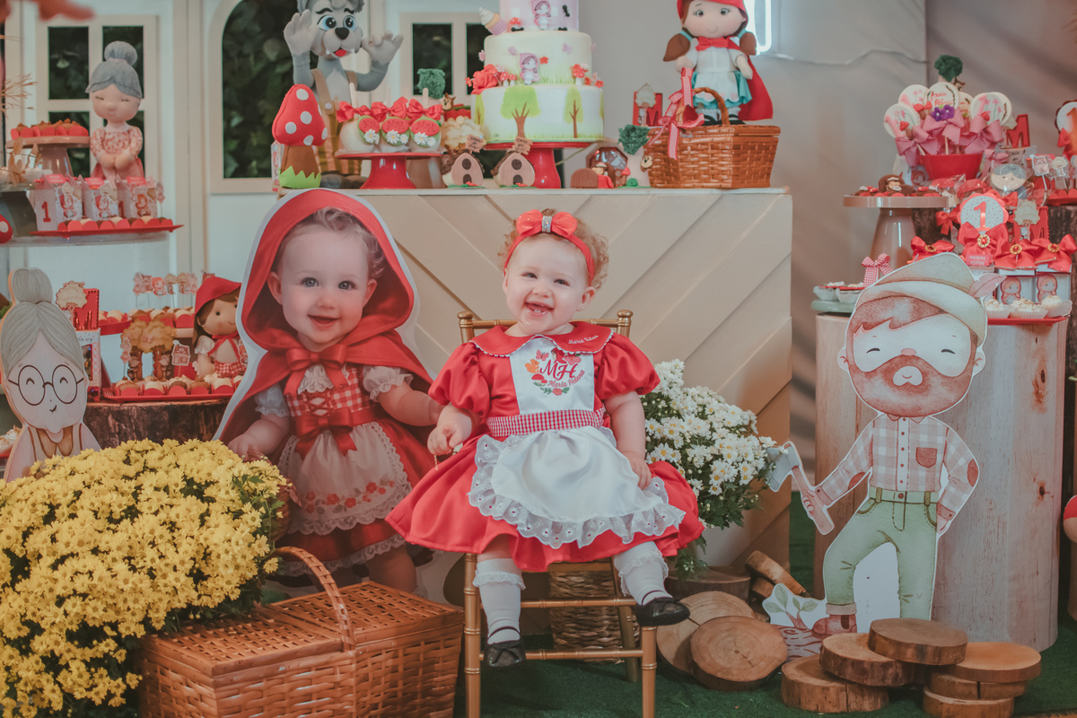 Uma bebês entada em uma cadeirinha, usando um vestido vermelho e em frente a uma decoração de tema chpauzinho vermelho, na festa de aniversário de 1 ano.
