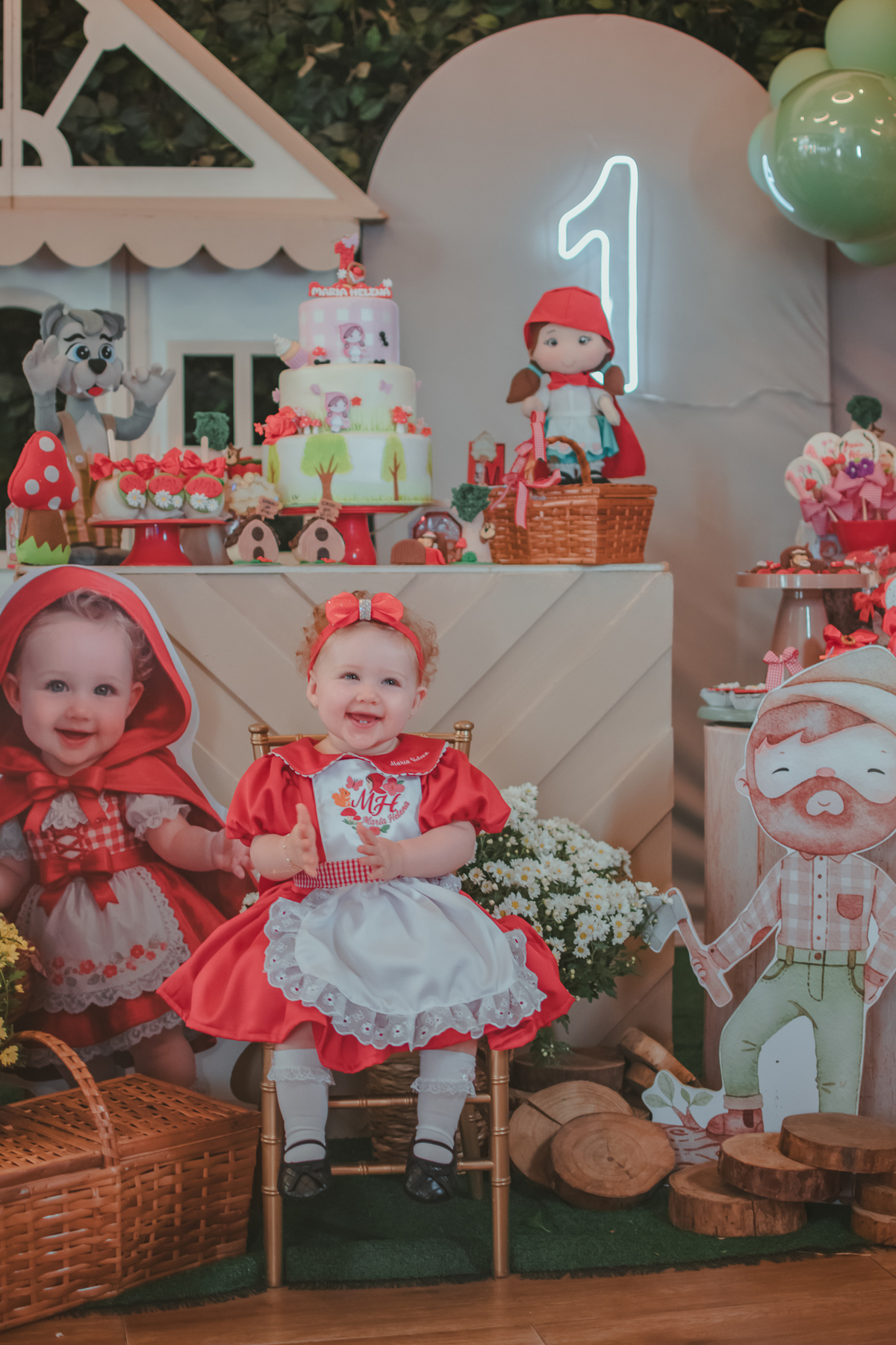 Uma bebês entada em uma cadeirinha, usando um vestido vermelho e em frente a uma decoração de tema chpauzinho vermelho, na festa de aniversário de 1 ano.