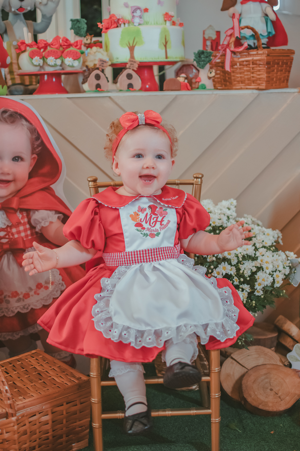 Uma bebês entada em uma cadeirinha, usando um vestido vermelho e em frente a uma decoração de tema chpauzinho vermelho, na festa de aniversário de 1 ano.