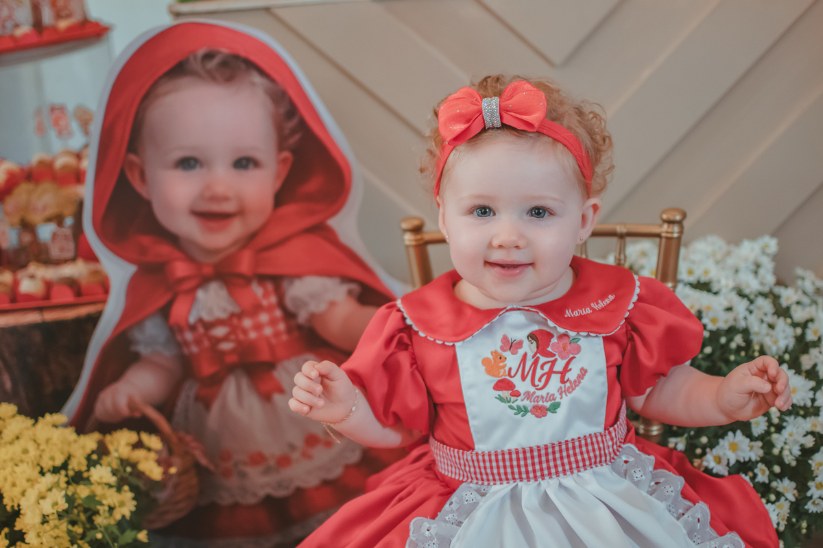 Uma bebês entada em uma cadeirinha, usando um vestido vermelho e em frente a uma decoração de tema chpauzinho vermelho, na festa de aniversário de 1 ano.