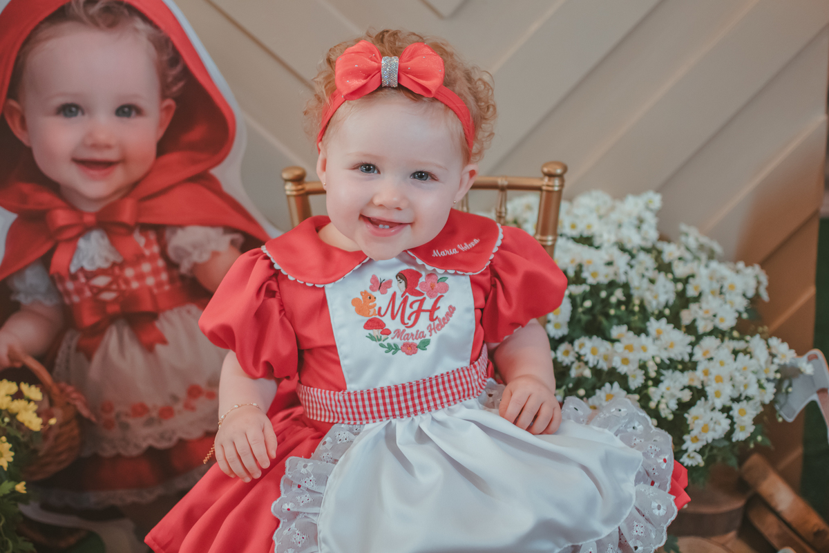 Uma bebês entada em uma cadeirinha, usando um vestido vermelho e em frente a uma decoração de tema chpauzinho vermelho, na festa de aniversário de 1 ano.