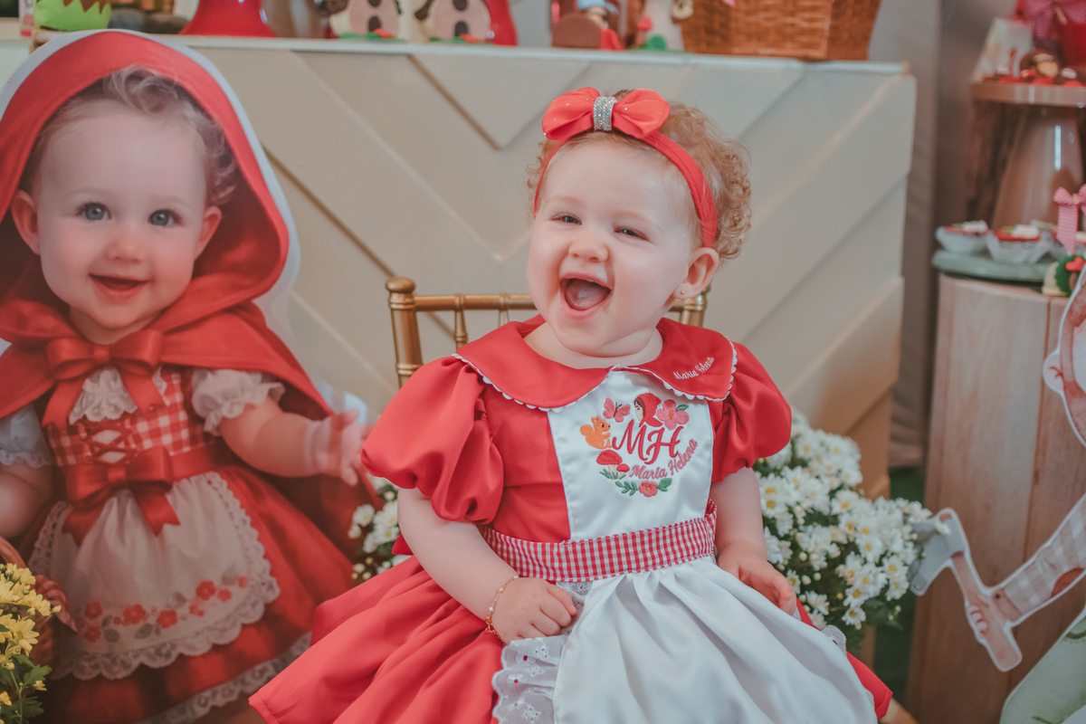 Uma bebês entada em uma cadeirinha, usando um vestido vermelho e em frente a uma decoração de tema chpauzinho vermelho, na festa de aniversário de 1 ano.
