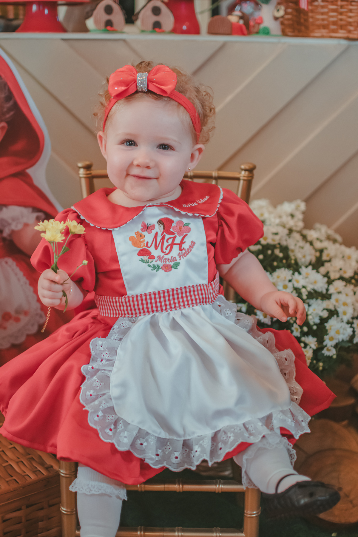 Uma bebês entada em uma cadeirinha, segurando uma flor amarela e usando um vestido vermelho e em frente a uma decoração de tema chpauzinho vermelho, na festa de aniversário de 1 ano.