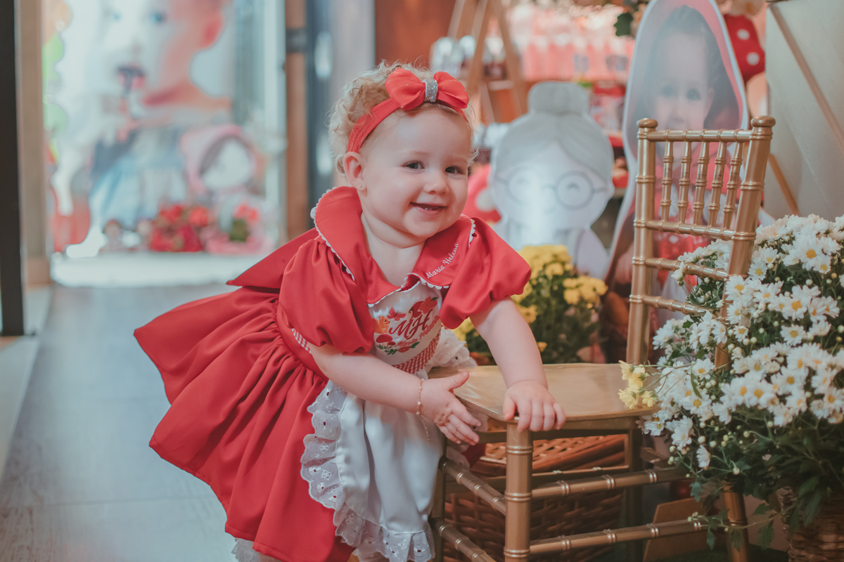 Uma bebês entada em uma cadeirinha, usando um vestido vermelho e em frente a uma decoração de tema chpauzinho vermelho, na festa de aniversário de 1 ano.
