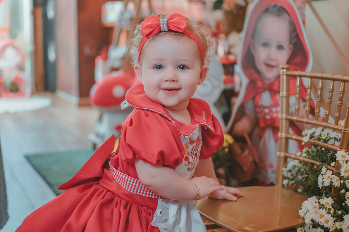 Uma bebês entada em uma cadeirinha, usando um vestido vermelho e em frente a uma decoração de tema chpauzinho vermelho, na festa de aniversário de 1 ano.