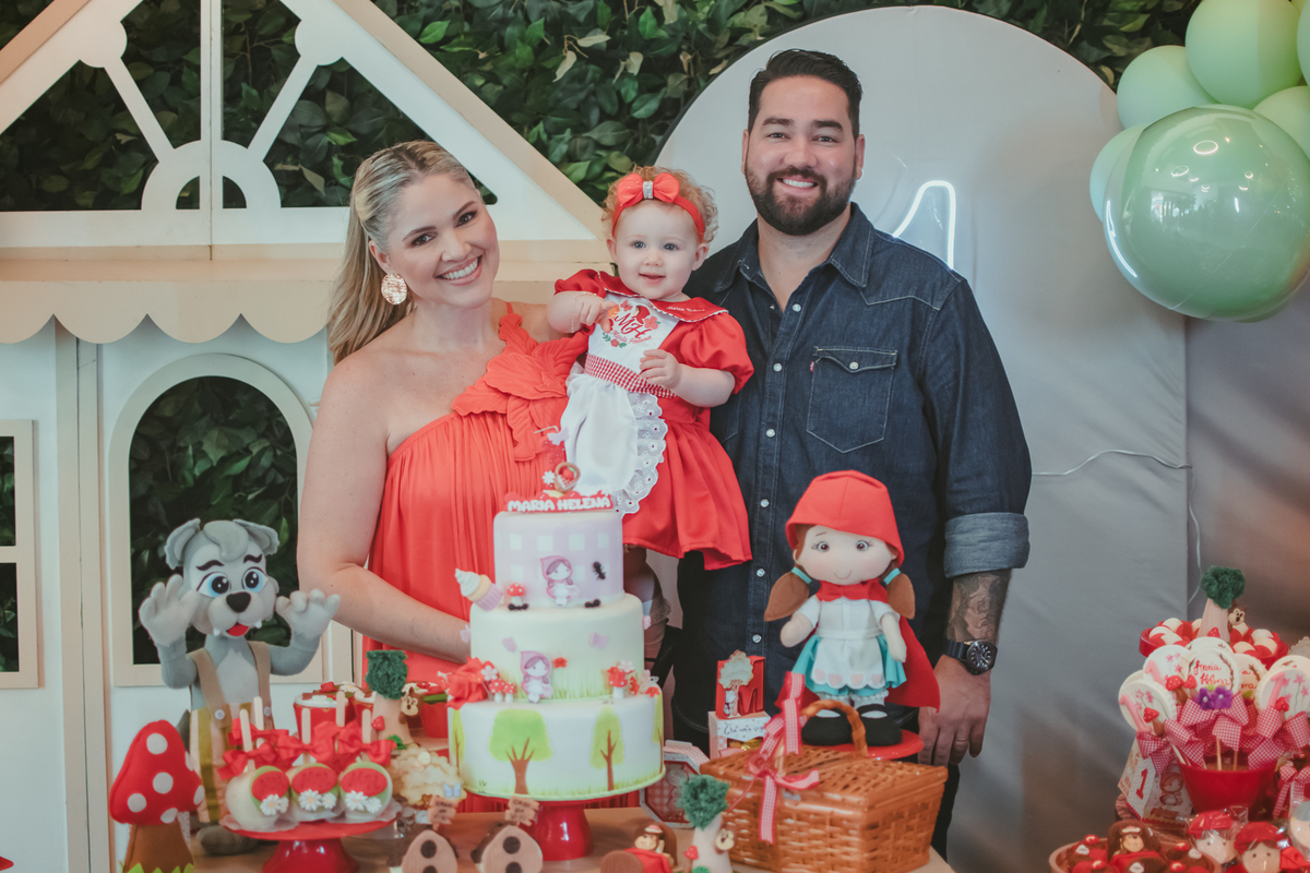 uma mulher de vestido vermelho e um homem de camisa azul segurando uma bebê de 1 aninho em frente uma decoração de chápeuzinho vermelho em uma festa de aniversário infantil.