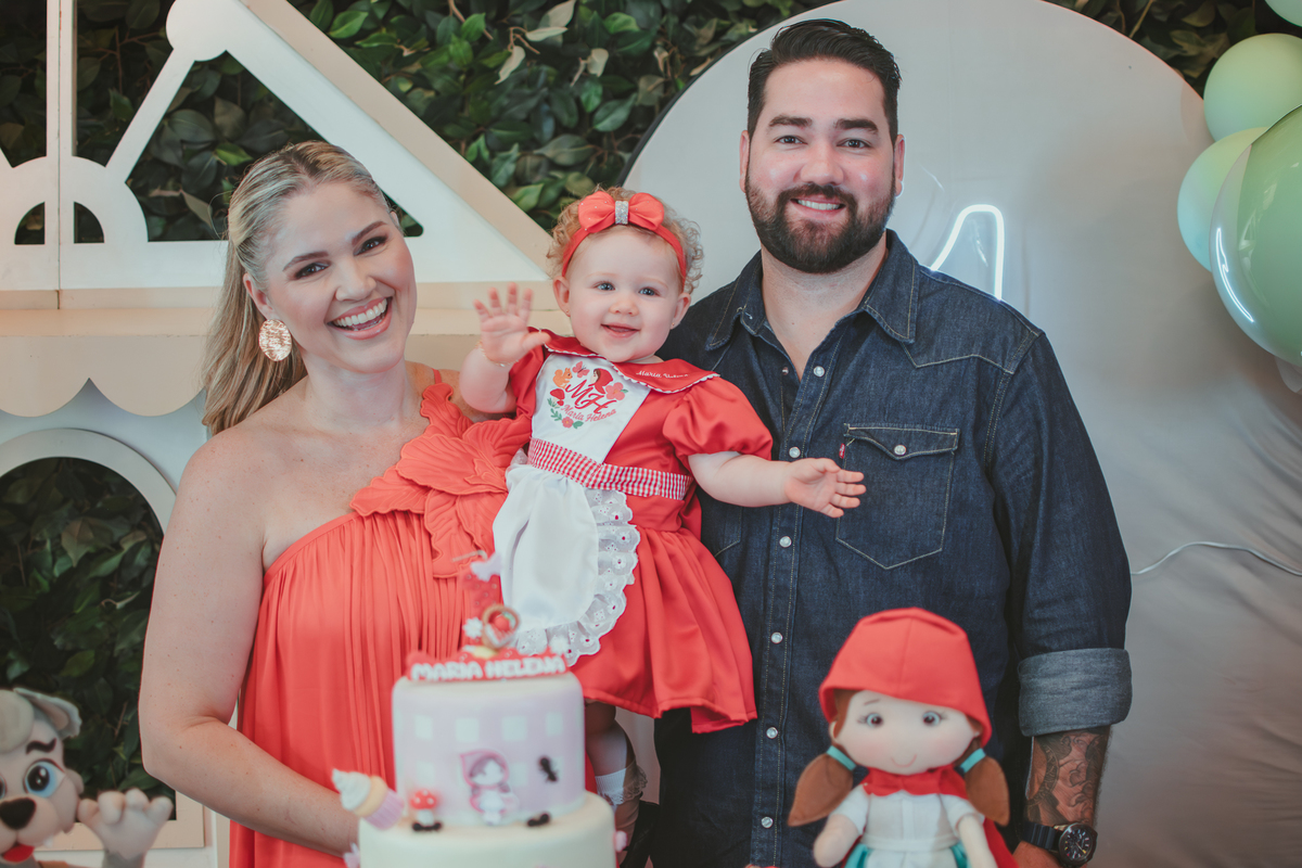 uma mulher de vestido vermelho e um homem de camisa azul segurando uma bebê de 1 aninho em frente uma decoração de chápeuzinho vermelho em uma festa de aniversário infantil.
